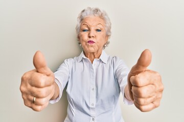 Senior grey-haired woman doing thumbs up positive gesture puffing cheeks with funny face. mouth inflated with air, catching air.