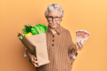 Senior grey-haired woman holding groceries and israel shekels banknotes skeptic and nervous,...
