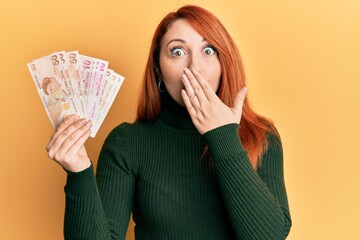 Beautiful redhead woman holding turkish lira banknotes covering mouth with hand, shocked and afraid for mistake. surprised expression