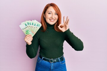 Beautiful redhead woman holding 50 hong kong dollars banknotes doing ok sign with fingers, smiling friendly gesturing excellent symbol