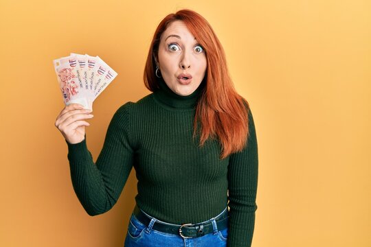 Beautiful redhead woman holding 10 singapore dollars banknotes scared and amazed with open mouth for surprise, disbelief face