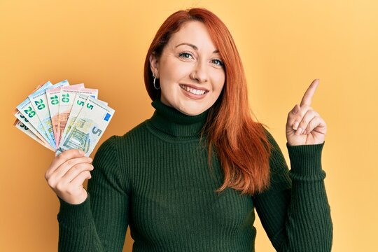 Beautiful redhead woman holding euro banknotes smiling happy pointing with hand and finger to the side