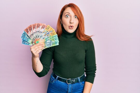 Beautiful redhead woman holding australian dollars scared and amazed with open mouth for surprise, disbelief face