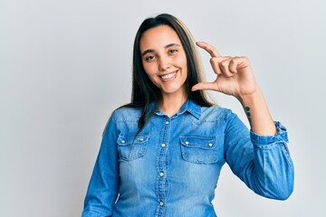 Young hispanic woman wearing casual denim jacket smiling and confident gesturing with hand doing small size sign with fingers looking and the camera. measure concept.