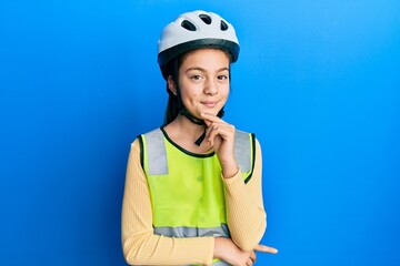 Beautiful brunette little girl wearing bike helmet and reflective vest looking confident at the camera with smile with crossed arms and hand raised on chin. thinking positive.