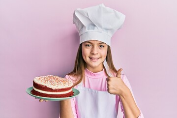 Beautiful brunette little girl wearing baker uniform holding homemade cake smiling happy and positive, thumb up doing excellent and approval sign