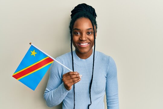 African american woman with braided hair holding democratic republic of the congo flag looking positive and happy standing and smiling with a confident smile showing teeth
