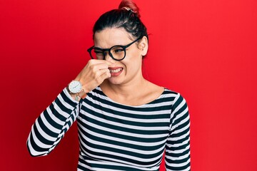 Young caucasian woman wearing casual clothes and glasses smelling something stinky and disgusting,...