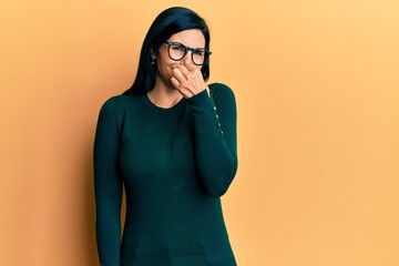 Young caucasian woman wearing casual clothes and glasses smelling something stinky and disgusting,...