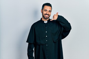 Handsome hispanic man with beard wearing catholic priest robe smiling doing phone gesture with hand and fingers like talking on the telephone. communicating concepts.