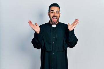 Handsome hispanic man with beard wearing catholic priest robe celebrating crazy and amazed for...