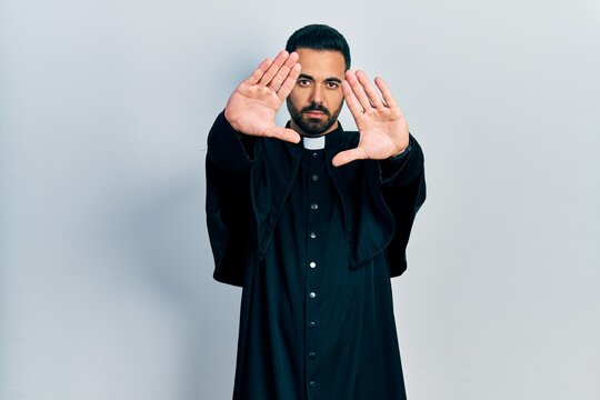 Handsome hispanic man with beard wearing catholic priest robe doing frame using hands palms and fingers, camera perspective