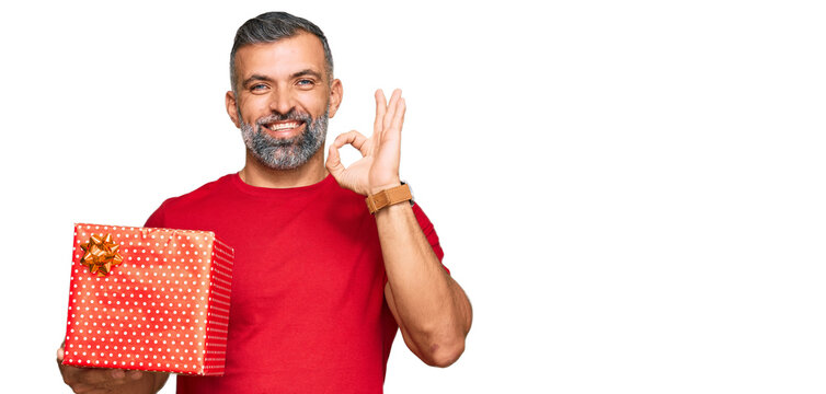 Middle age handsome man holding gift doing ok sign with fingers, smiling friendly gesturing excellent symbol
