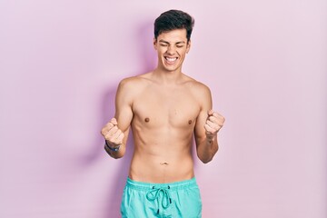 Fototapeta premium Young hispanic man wearing swimwear shirtless very happy and excited doing winner gesture with arms raised, smiling and screaming for success. celebration concept.