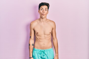 Fototapeta premium Young hispanic man wearing swimwear shirtless making fish face with lips, crazy and comical gesture. funny expression.