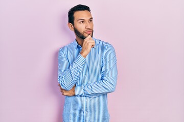 Hispanic man with beard wearing casual blue shirt with hand on chin thinking about question, pensive expression. smiling with thoughtful face. doubt concept.