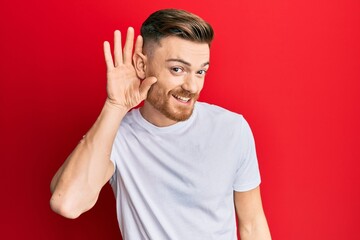 Fototapeta premium Young redhead man wearing casual white t shirt smiling with hand over ear listening and hearing to rumor or gossip. deafness concept.
