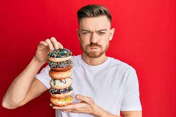 Young redhead man holding pile of tasty colorful doughnuts puffing cheeks with funny face. mouth...