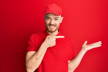 Young redhead man wearing delivery uniform and cap amazed and smiling to the camera while presenting with hand and pointing with finger.