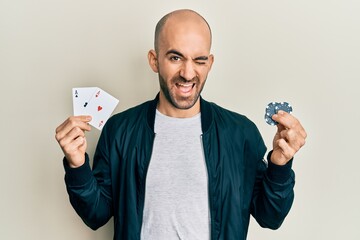Young hispanic man playing poker holding casino cards and chips winking looking at the camera with...