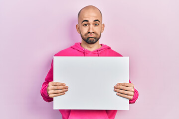 Young bald man holding blank empty banner puffing cheeks with funny face. mouth inflated with air, catching air.