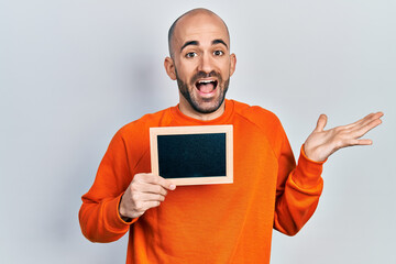 Young bald man holding blackboard celebrating achievement with happy smile and winner expression with raised hand