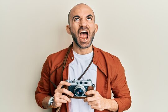 Young hispanic man holding vintage camera angry and mad screaming frustrated and furious, shouting with anger looking up.