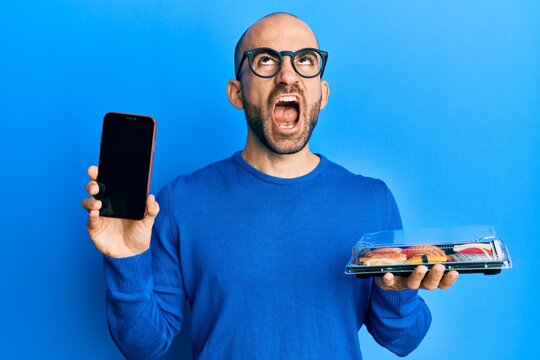 Young hispanic man holding take away food showing smartphone screen angry and mad screaming frustrated and furious, shouting with anger looking up.