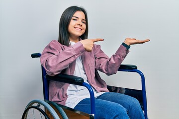 Young brunette woman sitting on wheelchair amazed and smiling to the camera while presenting with...