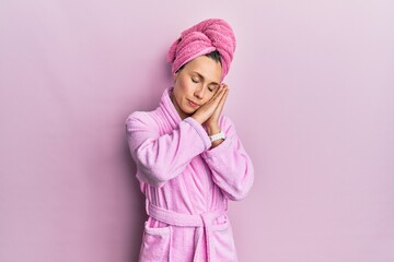 Young blonde woman wearing shower towel cap and bathrobe sleeping tired dreaming and posing with hands together while smiling with closed eyes. - obrazy, fototapety, plakaty