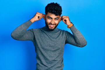 Arab man with beard wearing turtleneck sweater smiling pulling ears with fingers, funny gesture....