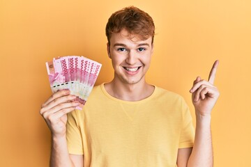 Young caucasian man holding 100000 indonesian rupiah smiling happy pointing with hand and finger to the side