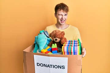 Young caucasian man holding donation box with toys winking looking at the camera with sexy...