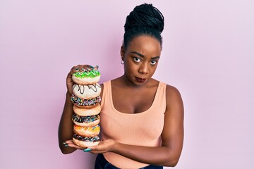 Young african american woman holding pile of tasty colorful doughnuts skeptic and nervous, frowning...