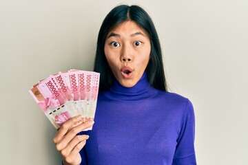 Young chinese woman holding indonesian rupiah banknotes scared and amazed with open mouth for...