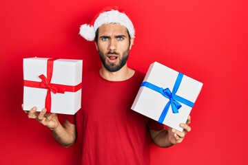 Young hispanic man wearing christmas hat and holding gifts in shock face, looking skeptical and...