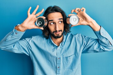 Young hispanic man holding canned food over eyes smiling looking to the side and staring away thinking.