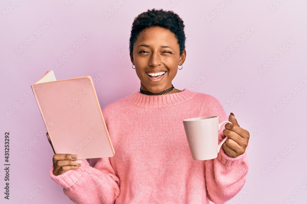 Wall mural Young african american girl reading a book and drinking a cup of coffee winking looking at the camera with sexy expression, cheerful and happy face.