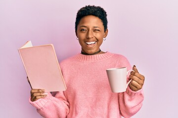 Young african american girl reading a book and drinking a cup of coffee winking looking at the...