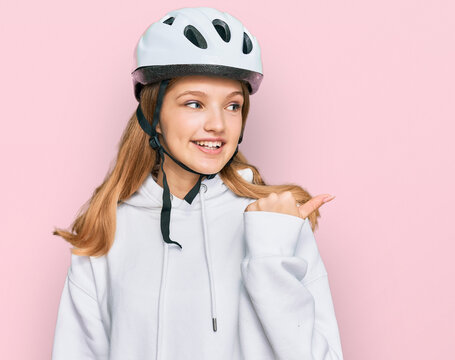 Beautiful young caucasian girl wearing bike helmet smiling with happy face looking and pointing to the side with thumb up.