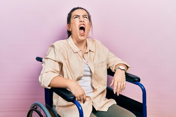 Young hispanic woman sitting on wheelchair angry and mad screaming frustrated and furious, shouting with anger. rage and aggressive concept.