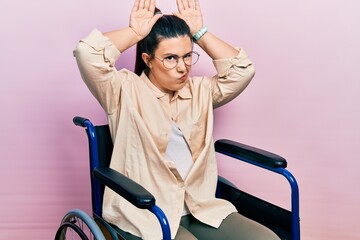 Young hispanic woman sitting on wheelchair doing bunny ears gesture with hands palms looking cynical and skeptical. easter rabbit concept.