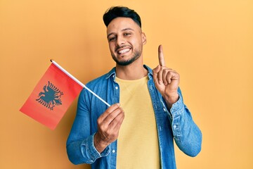 Young arab man holding albania flag smiling with an idea or question pointing finger with happy face, number one