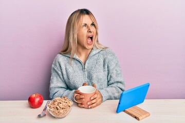 Young caucasian woman having breakfast looking at the tablet sitting on the tablet angry and mad screaming frustrated and furious, shouting with anger looking up.