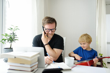 Fototapeta na wymiar Father freelancer working from home with laptop during quarantine. Naughty son distracts. Home office and parenthood at same time. Exhausted parent with hyperactive child.