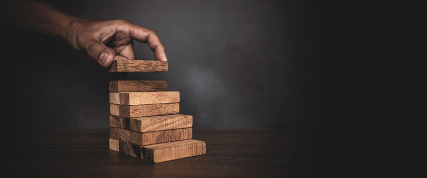 Close-up hand is placing wood block tower stacked in pyramid shape with caution to prevent collapse or crash concepts of financial risk management and strategic planning.