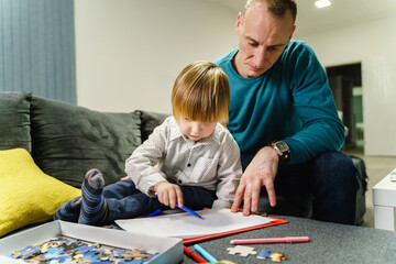Small caucasian boy sitting on the sofa bed with his dad playing with felt pen - Son and father spending time together learning and drawing at home - family time parenthood and childhood concept