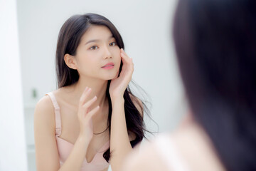 Beautiful young asian woman smiling look at mirror of checking face with skin care and cosmetic for rejuvenation and hygiene, beauty girl happy clean facial with cream or lotion and for health.