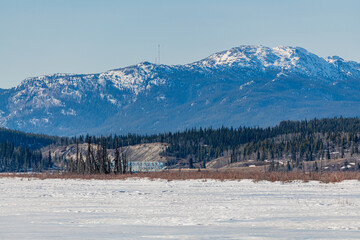 Fototapeta na wymiar Snow-capped mountain wilderness view in northern Canada with bright blue sky background in the melting, thawing season of Spring in April. Frozen Yukon river below. 