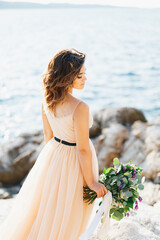 Fototapeta na wymiar Beautiful pensive bride stands turned sideways with a bouquet of flowers on a rock above the sea